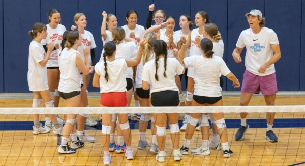 A group of teenage girls in sportswear stands in a circle, raising their hands in a cheer on a volleyball court, with an adult coach in shorts and a camp shirt joining in enthusiastically.