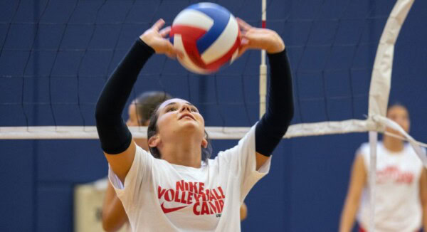 A volleyball player in a white Nike Volleyball Camp shirt prepares to set a volleyball during an indoor game. A net and another player in similar attire are visible in the background.