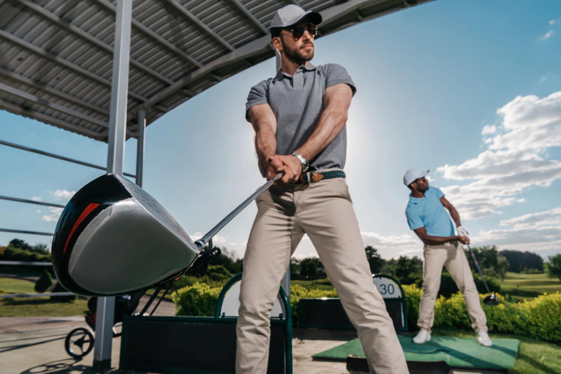 dynamic image capturing two golfers at a driving range, preparing to swing their clubs against a backdrop of blue skies and lush greenery.