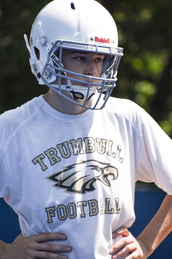 A football player wearing a white helmet and a Trumbull Football t-shirt stands with hands on hips, looking ahead with a focused expression. Trees and sunlight are visible in the background.